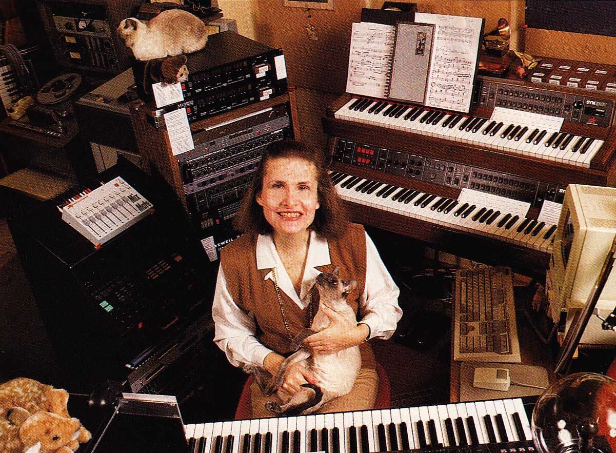 A photograph, apparently scanned from a magazine. Wendy Carlos, a white woman aged around 50 (in 1989), smiling up at the camera from the centre of a magnificent collection of music tech. She is holding a siamese cat, and another sits on top of a studio rack.